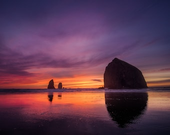 May include: A silhouette of Haystack Rock, a large rock formation, at sunset with a colorful sky and a reflection of the sky in the water.