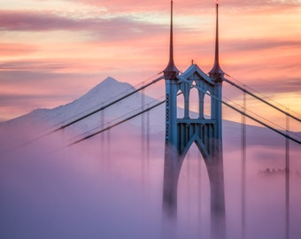 May include: A scenic photograph of a bridge shrouded in fog at sunrise. The bridge is teal and has two tall towers with spires. The sky is a mix of pink, orange, and purple hues. A mountain is visible in the background.