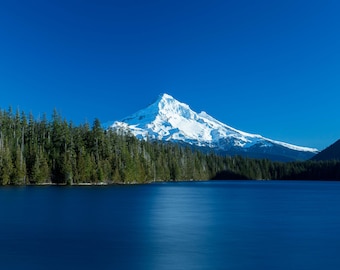 May include: A snow-capped mountain peak rises above a dark blue lake, with a lush forest of evergreen trees in the foreground.