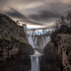 May include: A dramatic view of a waterfall cascading down a rocky cliff face. The water is flowing over multiple tiers, creating a misty spray. The sky is overcast with dark clouds, adding to the moody atmosphere.