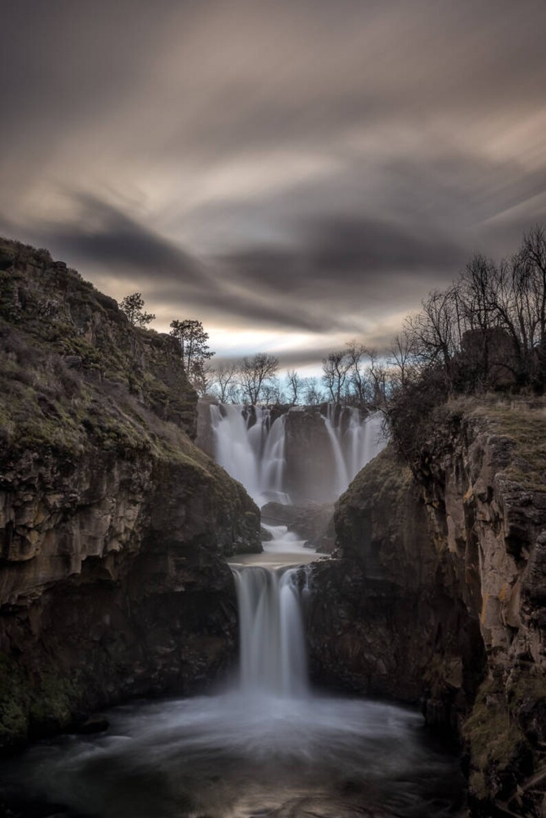 May include: A dramatic view of a waterfall cascading down a rocky cliff face. The water is flowing over multiple tiers, creating a misty spray. The sky is overcast with dark clouds, adding to the moody atmosphere.