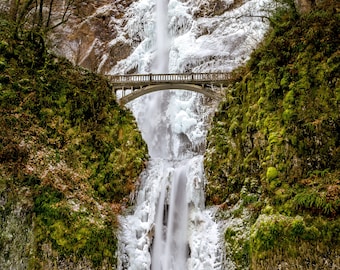 May include: A scenic photograph of a waterfall cascading down a rocky cliff face. A stone bridge spans the falls, surrounded by lush green moss and trees. The water is partially frozen, creating an icy effect.