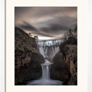 May include: A dramatic photograph of a waterfall cascading down a rocky cliff face. The water is flowing over multiple tiers, creating a misty spray. The sky above is a cloudy grey, with hints of blue peeking through.