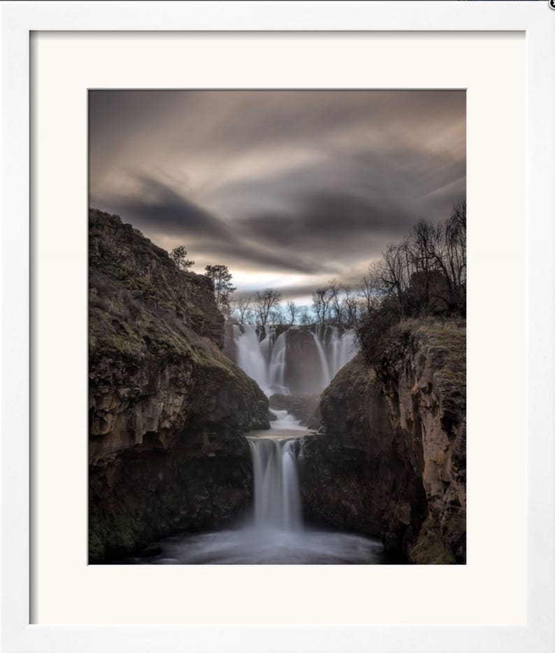May include: A dramatic photograph of a waterfall cascading down a rocky cliff face. The water is flowing over multiple tiers, creating a misty spray. The sky above is a cloudy grey, with hints of blue peeking through.