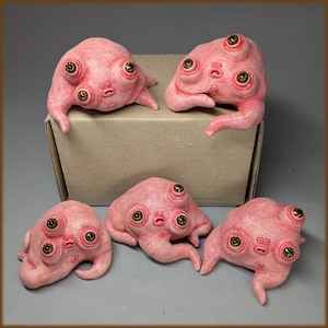 Peut inclure: Cinq petites sculptures roses, charnues et en forme de goutte, à la peau texturée, chacune dotée de trois yeux bruns réalistes et d'un minuscule visage à la bouche ouverte. Deux de ces créatures sont posées sur une boîte en carton beige.