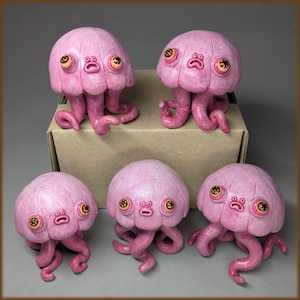 May include: Five small, pink, sculpted jellyfish figurines with rounded, segmented caps, multiple curling tentacles, and two prominent, wide-set eyes with golden irises. Each figure features a small, centred mouth with a surprised expression.