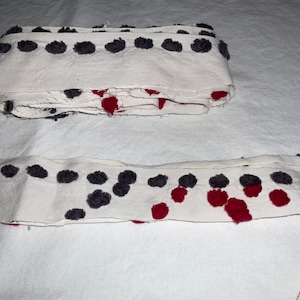 May include: White fabric trim with a row of gray and burgundy pom-poms. The trim is folded and laid out on a white surface.