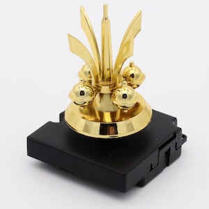 May include: A gold-colored clock mechanism with a decorative top featuring a central spire and four round ornaments. The mechanism is mounted on a black square base.