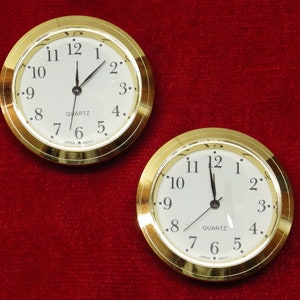 May include: Two gold-rimmed, round, white-faced quartz clocks with black hands. The clocks are set to 12:00 and have black numbers around the face. The text "QUARTZ" and "JAPAN MOVI" are printed on the face of each clock.