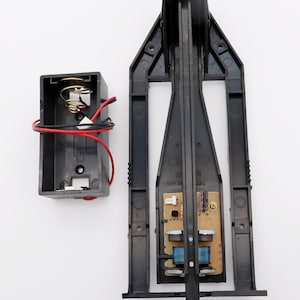 May include: A black battery compartment with a spring and red and black wires, and a black circuit board with a small circuit board attached to it. The circuit board has a small gold screw and two blue components. The text on the circuit board reads "(662-24) 21N".