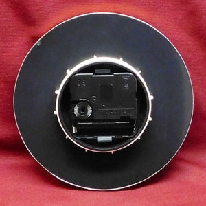 May include: A round clock back with a black face and a white rim. The clock mechanism is visible, featuring a black motor and a small silver gear. The clock is set against a red background.