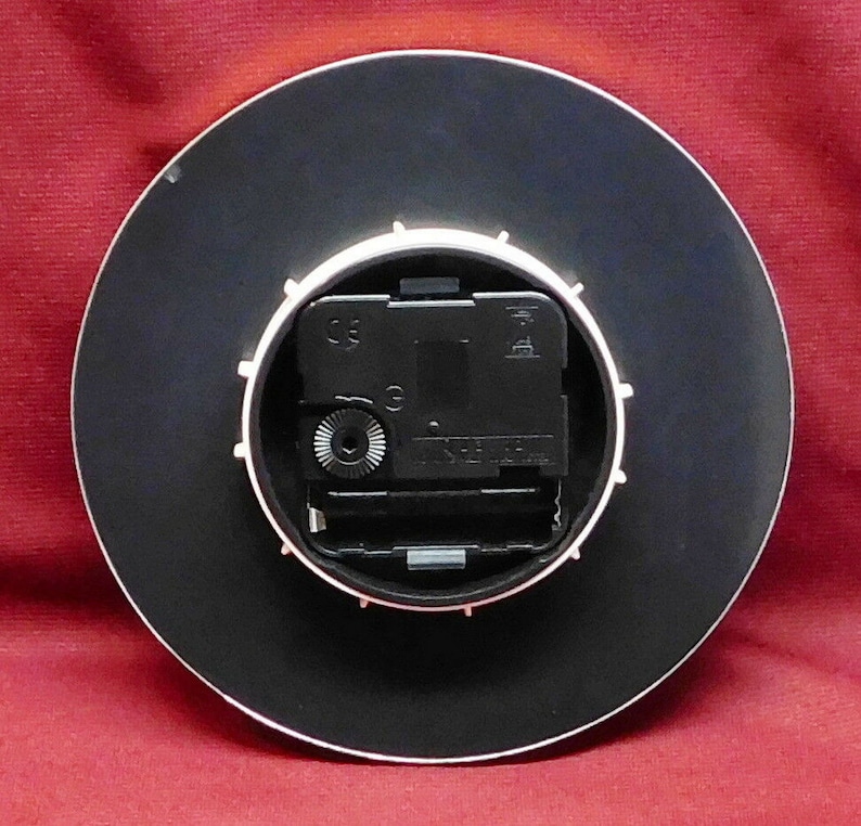 May include: A round clock back with a black face and a white rim. The clock mechanism is visible, featuring a black motor and a small silver gear. The clock is set against a red background.