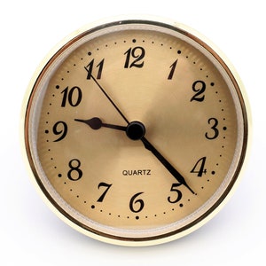 May include: A gold-colored clock face with black hands and black numbers. The clock face is surrounded by a gold-colored ring with a gold-colored bezel. The clock face has a white background with black markings. The clock face reads "QUARTZ".
