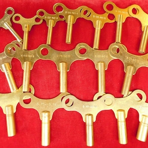 May include: A collection of 12 gold-toned clock winding keys with different numbers and letters printed on them. The keys are arranged in a random pattern on a red surface.