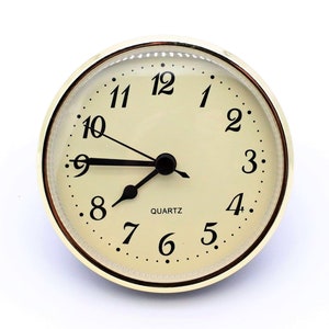 May include: A gold-colored clock face with black numbers and hands. The clock face is surrounded by a gold-colored ring. The clock face reads "QUARTZ".