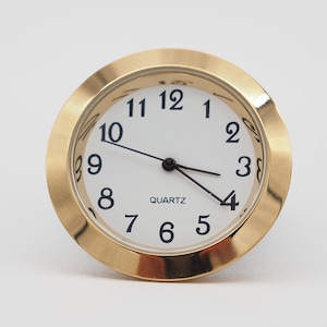 May include: A gold-colored clock face with black numbers and hands. The clock face is surrounded by a gold-colored bezel. The clock face reads "QUARTZ".