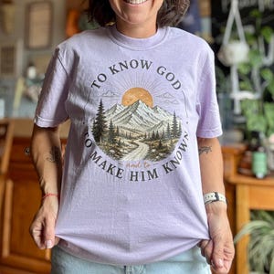 Può includere: Una t-shirt a maniche corte color viola chiaro con una grafica di un paesaggio montano con pini, un sentiero tortuoso e un sole. Il testo TO KNOW GOD AND TO MAKE HIM KNOWN è disposto in modo circolare attorno all'illustrazione centrale.