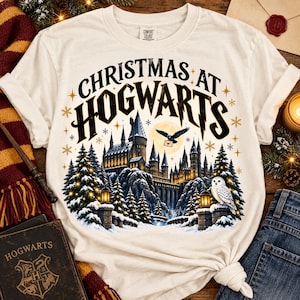 May include: A cream-colored short-sleeve t-shirt featuring a graphic print of a castle in a snowy landscape with pine trees, a flying owl carrying a letter, and the text "CHRISTMAS AT HOGWARTS" in a bold, dark, stylized font with gold stars.