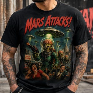 May include: A black t-shirt featuring a graphic print with the text MARS ATTACKS! in red. The illustration depicts a green alien with an exposed brain in a glass helmet holding a ray gun, with flying saucers, city buildings, and people.