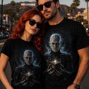 May include: A black short-sleeved t-shirt featuring a graphic print of a pale, pin-headed figure with a dark, textured torso holding a glowing cube. The design includes intricate circular patterns and blue energy effects surrounding the central character.