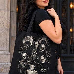 May include: A black tote bag featuring a detailed monochromatic graphic print of classic horror movie monsters, including a vampire, a mummy, a creature from the lagoon, a monster, and a bride, all set against a dark, moody background.