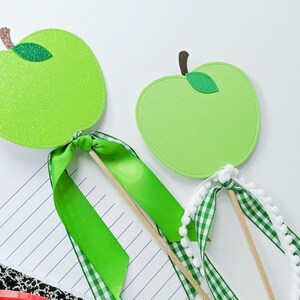 May include: Two green felt apple cutouts on wooden sticks with green and white ribbon bows. The apple on the left has glitter on it.
