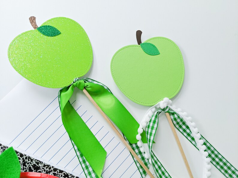 May include: Two green felt apple cutouts on wooden sticks with green and white ribbon bows. The apple on the left has glitter on it.