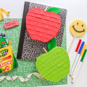 May include: A red and green apple-shaped pinata with a yellow smiley face lollipop, crayons, a measuring tape, and school supplies on a green and black alphabet chart.