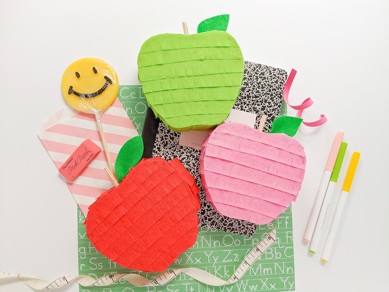 May include: Three apple-shaped pinatas, one red, one green, and one pink, are arranged on a green and black alphabet mat. A yellow smiley face lollipop, a pink eraser, and three colorful markers are also visible.