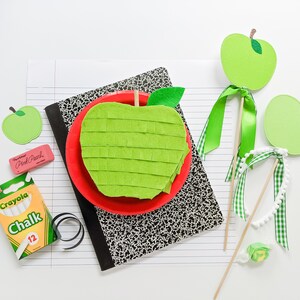 May include: A green felt apple pinata with a red paper plate and a green felt leaf. There are two green felt apple decorations on sticks with green and white gingham ribbon. There is a box of Crayola chalk and a pink eraser with the text "Pink Pearl" on it.
