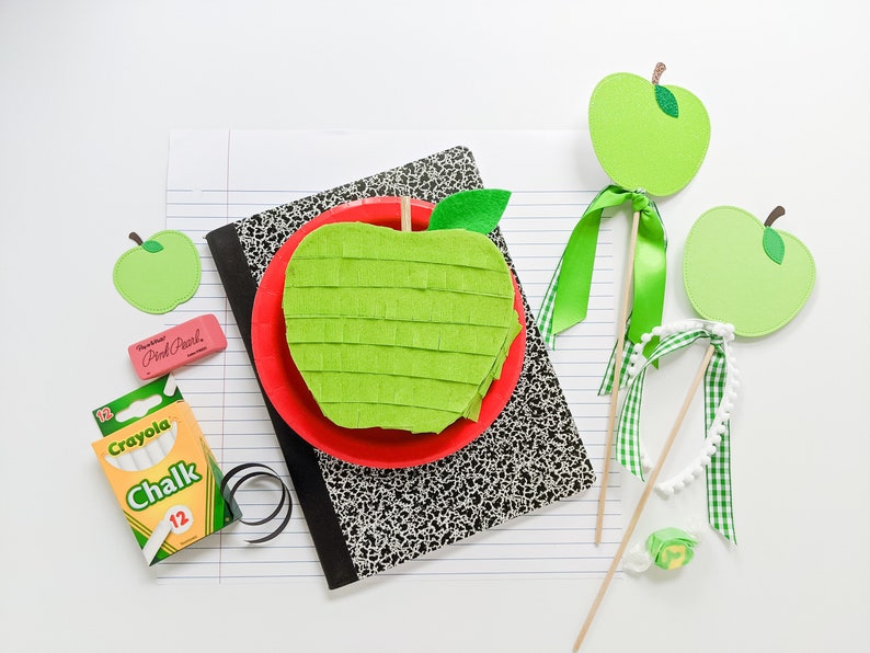 May include: A green felt apple pinata with a red paper plate and a green felt leaf. There are two green felt apple decorations on sticks with green and white gingham ribbon. There is a box of Crayola chalk and a pink eraser with the text "Pink Pearl" on it.
