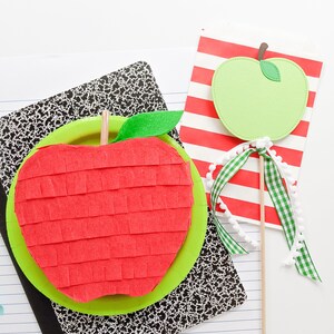 May include: A red and green apple shaped pinata with a green leaf on top. The pinata is sitting on a black and white notebook. A green apple on a stick is in the background.