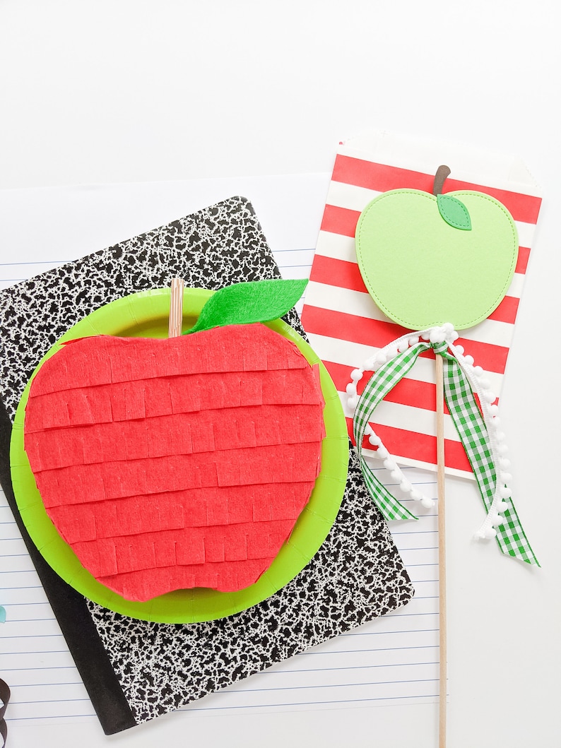 May include: A red and green apple shaped pinata with a green leaf on top. The pinata is sitting on a black and white notebook. A green apple on a stick is in the background.