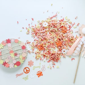 May include: A pink and orange paper flower pinata with a peace sign, groovy, and dance text. The pinata is surrounded by pink and orange confetti and a pink and white wand with white flowers.