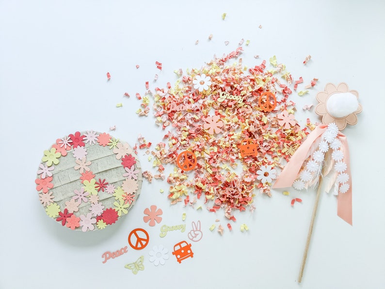 May include: A pink and orange paper flower pinata with a peace sign, groovy, and dance text. The pinata is surrounded by pink and orange confetti and a pink and white wand with white flowers.