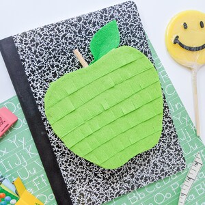 May include: A green apple-shaped pinata with a felt leaf on top, sitting on a black and white composition notebook. There are crayons, a pink eraser, and a green alphabet chart in the background.