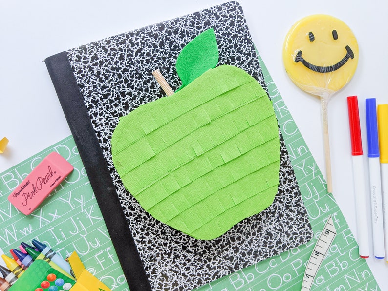 May include: A green apple-shaped pinata with a felt leaf on top, sitting on a black and white composition notebook. There are crayons, a pink eraser, and a green alphabet chart in the background.