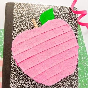 May include: A pink apple-shaped pi&ntilde;ata with a green felt leaf on top, sitting on a black and white speckled notebook.