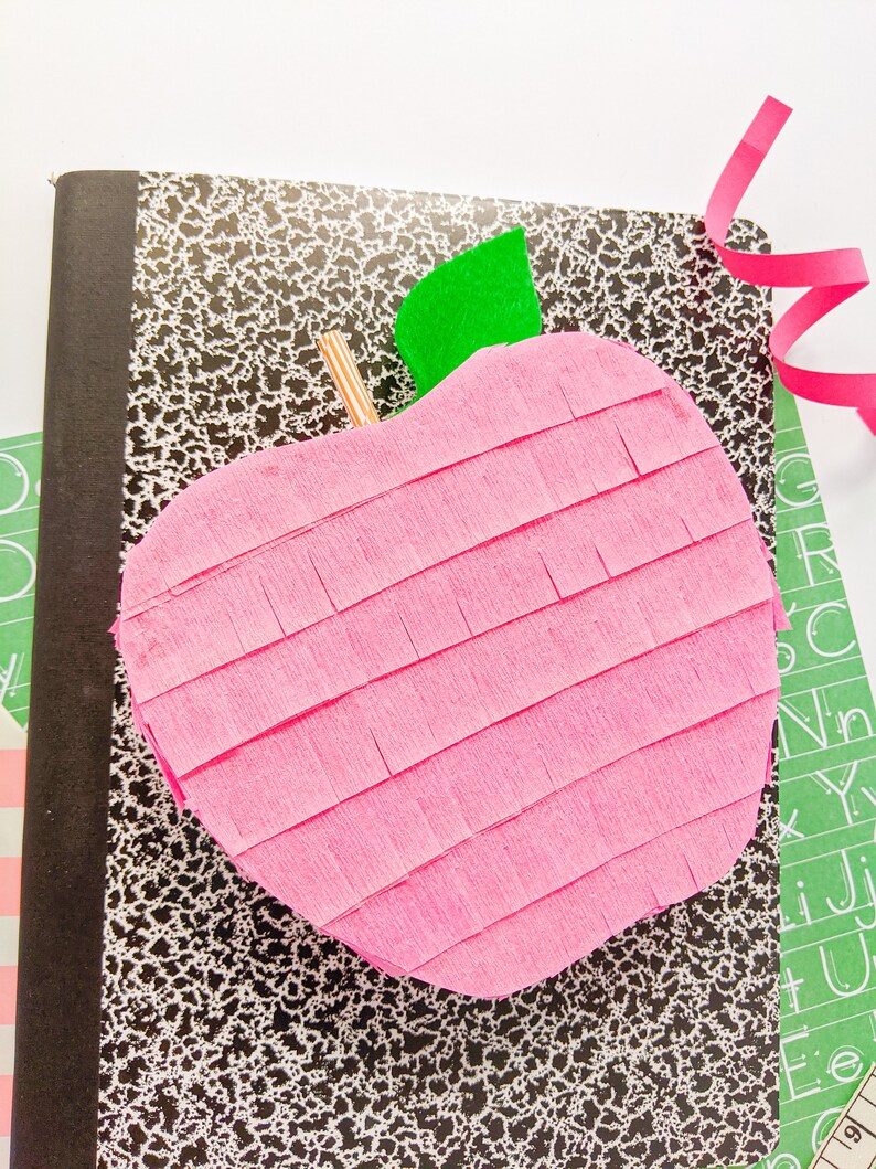May include: A pink apple-shaped pi&ntilde;ata with a green felt leaf on top, sitting on a black and white speckled notebook.