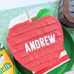 May include: A red apple-shaped pinata with the name "ANDREW" written on it. The pinata is decorated with green tissue paper and is sitting on a green plate.