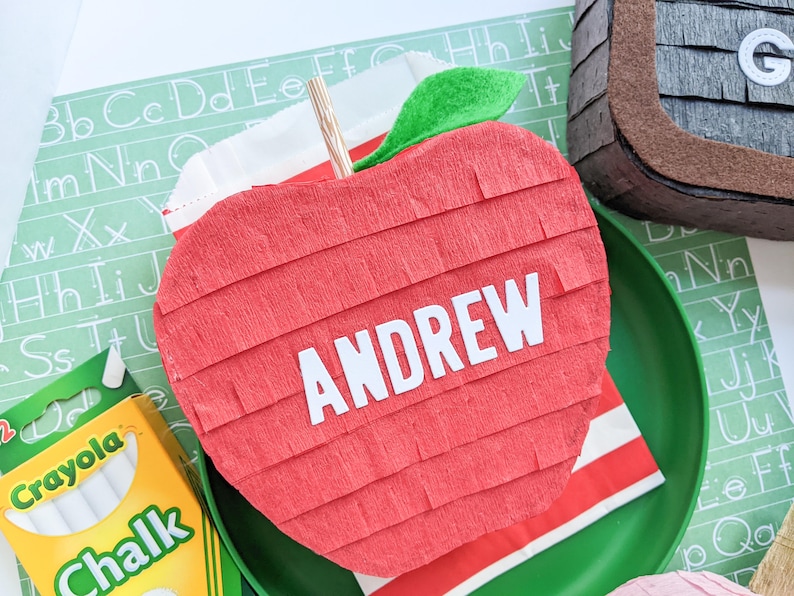 May include: A red apple-shaped pinata with the name "ANDREW" written on it. The pinata is decorated with green tissue paper and is sitting on a green plate.