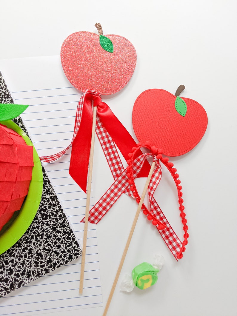 May include: Two red apple-shaped craft sticks with green leaves and red and white checkered ribbon. One apple has glitter and the other is plain. The sticks are on a white surface with a notebook and a green and yellow candy.