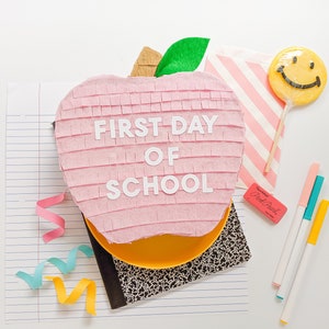May include: A pink apple-shaped pinata with the words "FIRST DAY OF SCHOOL" written on it. The pinata is surrounded by school supplies, including a notebook, pencils, and a smiley face lollipop.