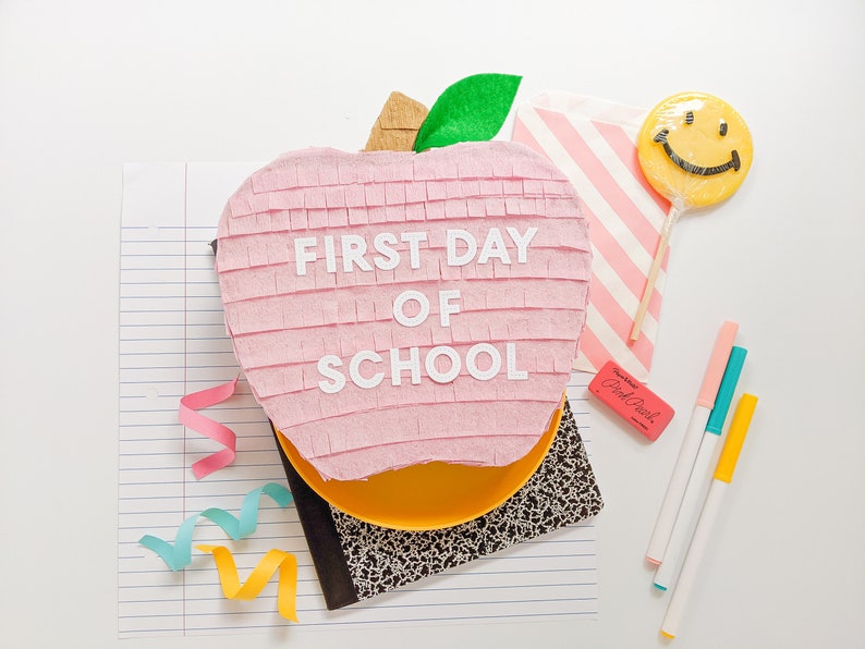 May include: A pink apple-shaped pinata with the words "FIRST DAY OF SCHOOL" written on it. The pinata is surrounded by school supplies, including a notebook, pencils, and a smiley face lollipop.