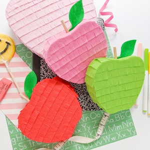 May include: Three apple-shaped pinatas, one pink, one red, and one green, are arranged on a green and white alphabet patterned surface. A yellow smiley face lollipop and a pink eraser are also visible.
