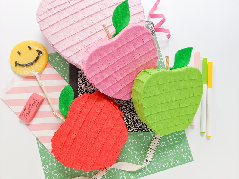 May include: Three apple-shaped pinatas, one pink, one red, and one green, are arranged on a green and white alphabet patterned surface. A yellow smiley face lollipop and a pink eraser are also visible.