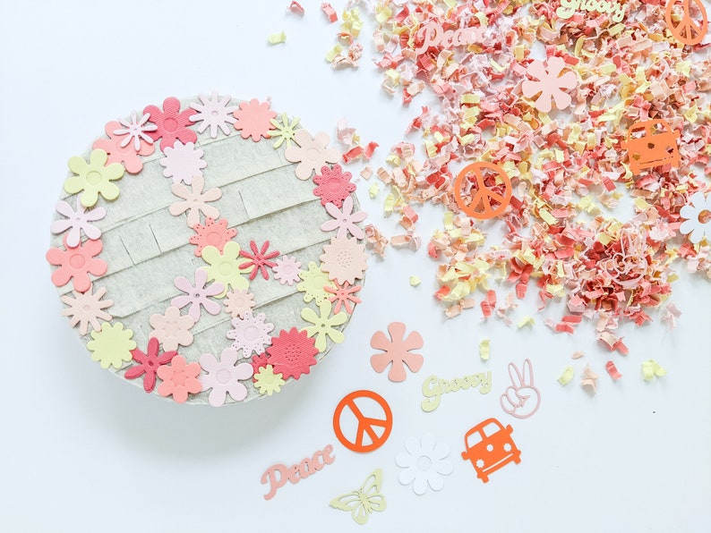 May include: A peace sign made of felt with pink, yellow, and orange flowers around the edges. The peace sign is surrounded by confetti and other paper cutouts, including the words "Peace" and "Groovy", a peace sign, a butterfly, and a car.