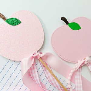 May include: Two pink apple-shaped wands with green leaves and pink and white gingham ribbon. The wands are on a white surface with a sheet of lined paper.