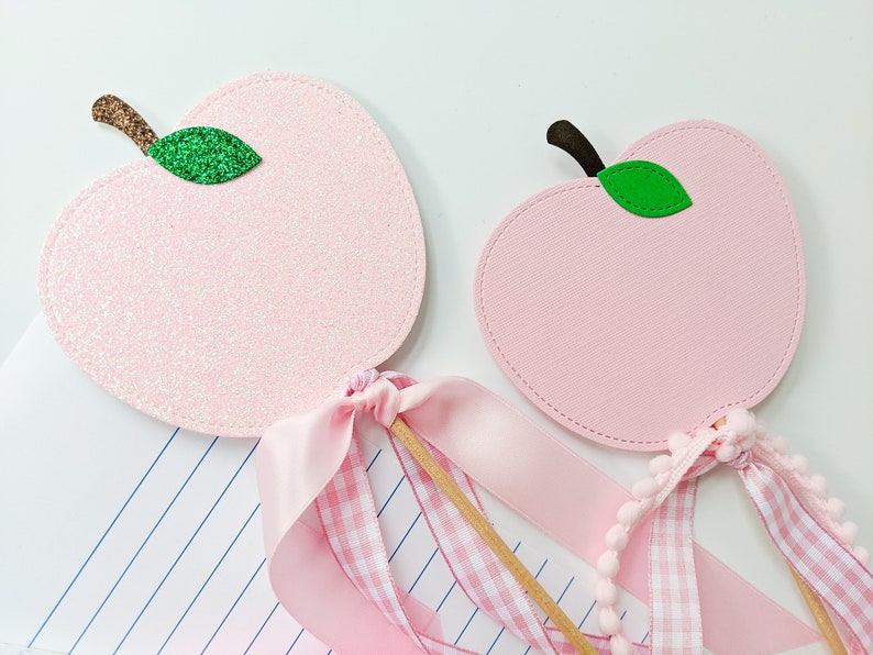 May include: Two pink apple-shaped wands with green leaves and pink and white gingham ribbon. The wands are on a white surface with a sheet of lined paper.