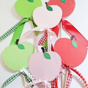 May include: Five apple-shaped paper crafts with glitter and ribbon accents. The apples are green, pink, and red. The ribbons are green and white gingham, pink and white gingham, and red and white gingham.